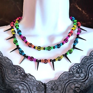 Puede incluir: Dos collares en capas con un esquema de colores arcoíris. Los collares presentan cuentas negras con caras sonrientes y detalles de púas negras. Los collares se exhiben en un maniquí blanco.