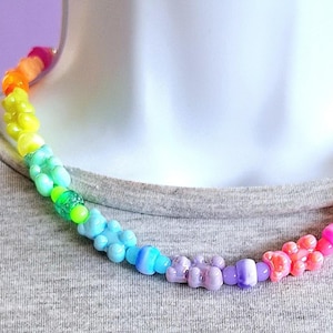 Könnte beinhalten: Eine farbenfrohe Halskette mit Gummibärchen-förmigen Perlen in einem Regenbogenverlauf. Die Perlen wechseln von Rosa zu Orange, Gelb, Grün, Blau und Lila. Die Halskette wird auf einem grauen T-Shirt getragen.