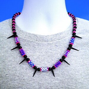 Puede incluir: Un collar de cuentas negro, rosa y azul con púas negras. El collar presenta secciones alternas de cuentas negras, cuentas estampadas rosas y azules y púas negras. El collar se muestra en un maniquí.