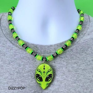 May include: A necklace featuring a lime green alien head pendant with black details and the text DIZZYPOP. The chain consists of alternating translucent lime green round beads and small black cylindrical spacers with silver-toned metallic accents.
