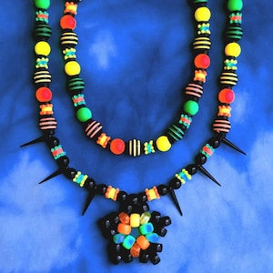 Puede incluir: Un collar de doble hebra con cuentas negras, verde neón, amarillas y naranjas. La hebra inferior incluye púas de plástico negro y un colgante central en forma de flor hecho de cuentas de plástico translúcidas y sólidas de colores.