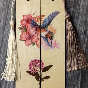 Può includere: Due segnalibri in legno con un colibrì e un motivo floreale. Il colibrì ha ali blu e un lungo becco, circondato da fiori rosa e pesca. Ogni segnalibro ha un nappa, uno color crema e uno marrone.