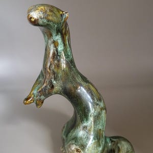 Könnte beinhalten: Keramikskulptur eines Tieres mit langem Hals und gebogenem Körper. Die Skulptur ist in Grün-, Braun- und Goldtönen glasiert. Das Tier sitzt mit erhobenen Armen. Die Skulptur hat eine glänzende Oberfläche.