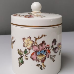 Könnte beinhalten: Ein weißes Keramikgefäß mit Deckel, verziert mit Blumen- und Vogelmotiven in Rosa, Gelb, Blau und Grün. Das Gefäß hat einen goldenen Rand und einen kleinen, runden Knopf oben. Das Design erinnert an Vintage-Porzellan.