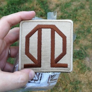 May include: Square patch with a tan background and a brown geometric design. The patch is held in a hand, and the background is blurred green grass.