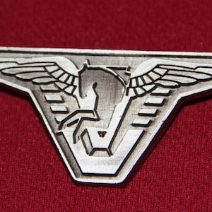 May include: A silver metal badge with a winged horse design. The horse is facing right and has a stylized wing on each side. The badge is shaped like a triangle with rounded corners.