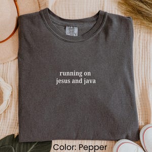 May include: Pepper-colored t-shirt with the phrase "running on jesus and java" in white. The shirt is displayed on a neutral-toned surface, with a hat and shoes visible in the background.