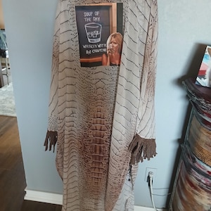 May include: A long, flowing kimono-style garment with a reptile skin pattern in shades of brown and beige. The garment has brown fringe on the sleeves and a small sign that says "Soup of the Day: Whiskey with H2O Croutons."