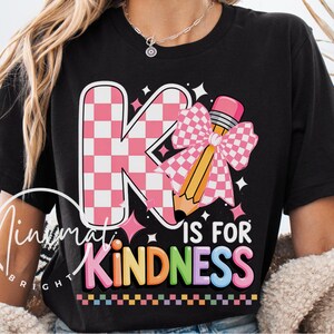 May include: Black t-shirt with a colorful graphic that says "K is for Kindness." The design features a pink and white checkered "K" with a pencil and bow. The text "Kindness" is in rainbow colors.