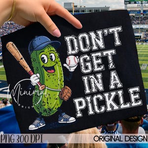 May include: Black t-shirt featuring a cartoon pickle character in a baseball uniform, holding a bat and baseball. The text "DON'T GET IN A PICKLE" is printed in white. The design includes a baseball stadium background.