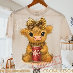May include: Cream-colored t-shirt featuring a cartoon cow with a leopard print bow, scarf, and a Dr. Pepper can. The cow is light brown with large eyes and a pink nose. The shirt has the words "Rustic Charm" and "Original Design" printed on it.