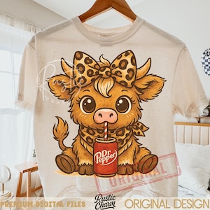 May include: Cream-colored t-shirt featuring a cartoon cow with a leopard print bow, bandana, and a Dr. Pepper can with a straw. The cow is brown with large eyes. The shirt is hanging on a wooden hanger.