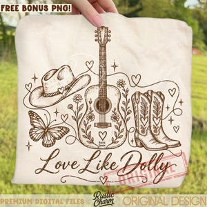 Puede incluir: Una camiseta color crema con un gráfico de líneas marrones de un sombrero de vaquero, una guitarra acústica, una mariposa y botas de vaquero, decorado con flores, corazones y estrellas. El texto dice Love Like Dolly y Rustic Charm.