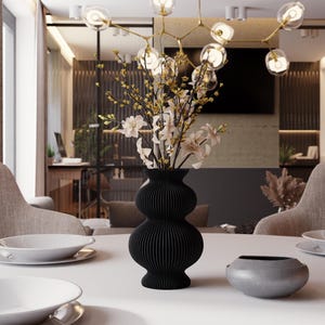 Op de afbeelding: Een zwarte, geribbelde vaas met een uniek, gelaagd ontwerp bevat een bloemstuk van witte en gele bloemen. De vaas staat op een witte tafel, met een kleine grijze kom en witte borden zichtbaar.