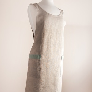 May include: A beige linen apron with a green trim and a pocket on the front. The apron is on a mannequin.