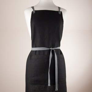 May include: A black linen apron with a large pocket and adjustable straps. The straps are grey and have a button closure. The apron is tied at the waist with a grey ribbon.