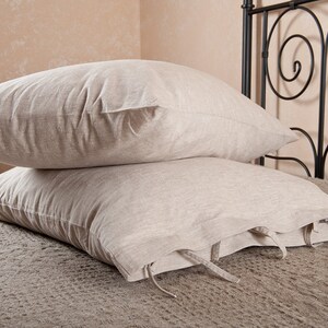 May include: Two beige linen pillowcases with ties, stacked on a bed with a black metal headboard.