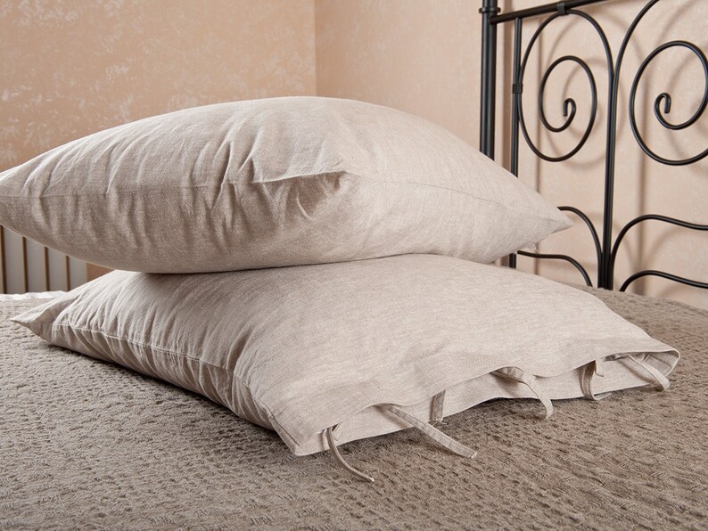May include: Two beige linen pillowcases with ties, stacked on a bed with a black metal headboard.