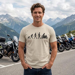 Könnte beinhalten: Ein beiges Kurzarm-T-Shirt mit einem schwarzen Grafikdruck, der die evolutionäre Entwicklung vom Primaten zum Menschen darstellt und mit einer Person auf einem Motorrad endet. Das Motiv ist mittig auf der Brust platziert.