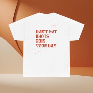 Könnte beinhalten: Weißes T-Shirt mit dem Text "DON'T LET IDIOTS RUIN YOUR DAY" in Rotbraun. Der Text wird von Sternen-Grafiken begleitet. Das T-Shirt hat einen Rundhalsausschnitt und kurze Ärmel, perfekt für den Alltag.