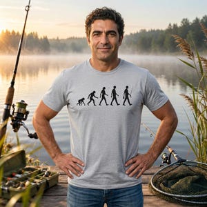 Könnte beinhalten: Ein hellgraues Kurzarm-T-Shirt mit einem schwarzen Grafikdruck der menschlichen Evolution, der mit einer Figur endet, die eine Angelrute hält. Das T-Shirt hat einen Rundhalsausschnitt und eine Standardpassform aus weichem, meliertem Stoff.
