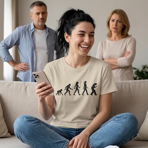 Könnte beinhalten: Ein beiges Kurzarm-T-Shirt mit einem schwarzen Grafikdruck, der die menschliche Evolutionssequenz darstellt und mit einer gebeugten Figur endet, die auf ein Smartphone schaut. Das T-Shirt hat einen Rundhalsausschnitt und eine Standardpassform.