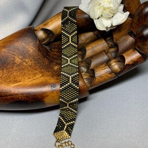 Könnte beinhalten: Ein schwarzes und goldenes Armband mit geometrischem Muster. Das Armband besteht aus kleinen, quadratischen Perlen und hat einen goldenen Kettenverschluss.