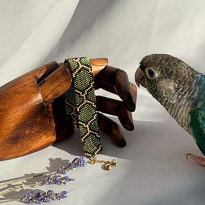 May include: A green and black beaded bracelet with a gold chain and charm. The bracelet is on a wooden hand model, and a green and blue parrot is looking at it.