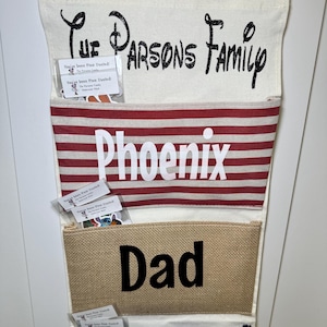 May include: A multi-pocket organizer with the text "The Parsons Family" in black script. The pockets are labeled "Phoenix", "Dad", and "Mom". The top pocket is beige, the second has red and white stripes, the third is burlap, and the bottom is navy blue.