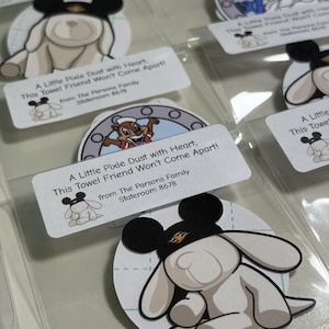 May include: Multiple circular stickers in clear plastic sleeves featuring cartoon characters like a beige rabbit wearing black mouse ears and a chipmunk in a porthole. Each sticker has a white label with the text A Little Pixie Dust with Heart, This Towel Friend Won't Come Apart! from The Parsons Family Stateroom 8678.