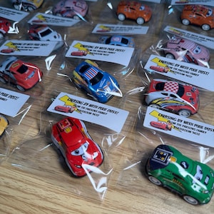 May include: A collection of toy race cars in individual clear plastic bags. The cars are various colors, including red, blue, pink, and orange, with racing-themed designs. Each bag includes a small white tag with text.