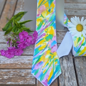 Puede incluir: Una corbata con base blanca y un patrón abstracto pintado con pinceladas amarillas, rosas y azul claro. La corbata se muestra junto a una pequeña margarita blanca con centro amarillo y un racimo de pequeñas flores de flox de color púrpura.