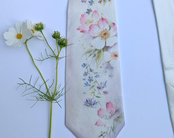May include: A white necktie with a watercolor floral design featuring pink, blue, and green flowers and leaves. The tie has a light pink gradient at the bottom. A white flower and stem are next to the tie.