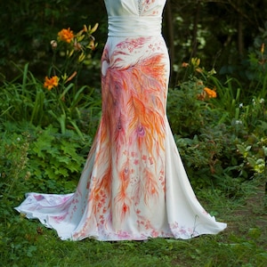 May include: A floor-length white sleeveless dress featuring a V-neckline with silver embellishments, a wide waistband, and a hand-painted design of orange and red peacock feathers and small floral accents extending from the hip to the hem.
