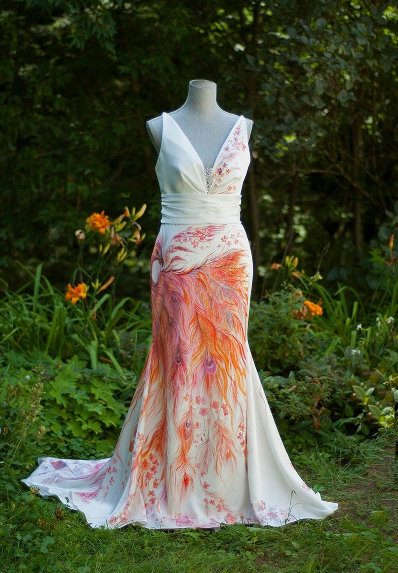 May include: A floor-length white sleeveless dress featuring a V-neckline with silver embellishments, a wide waistband, and a hand-painted design of orange and red peacock feathers and small floral accents extending from the hip to the hem.