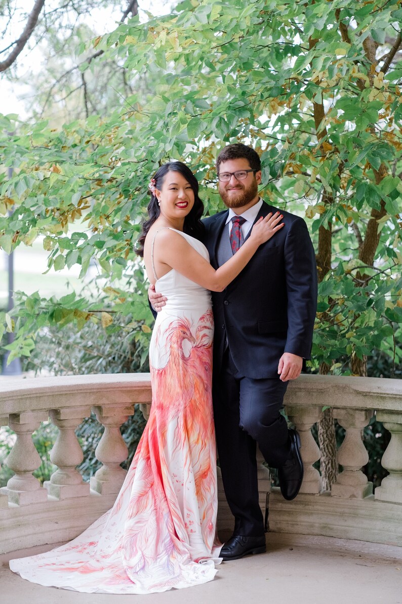 May include: A floor-length white gown featuring a hand-painted phoenix design in shades of red, orange, and purple, paired with a dark charcoal suit, a grey vest, a white dress shirt, and a red patterned necktie with a dark blazer and trousers.