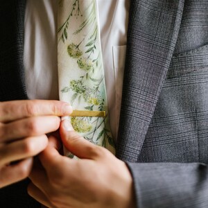 Puede incluir: Un hombre ajusta un pasador de corbata dorado en una corbata con estampado floral. La corbata es blanca con dise&ntilde;os florales verdes y amarillos. El hombre lleva un traje gris.