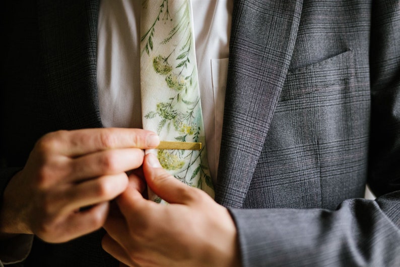 Puede incluir: Un hombre ajusta un pasador de corbata dorado en una corbata con estampado floral. La corbata es blanca con dise&ntilde;os florales verdes y amarillos. El hombre lleva un traje gris.
