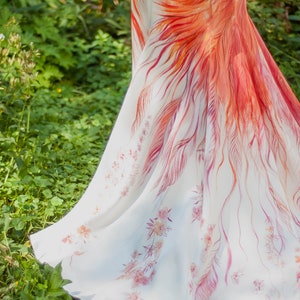 May include: A floor-length white skirt featuring a vibrant hand-painted design of large, sweeping orange and red feathers that cascade down the side, complemented by delicate, smaller pink floral patterns scattered across the lower hem area.