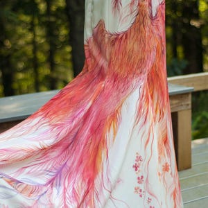 May include: A floor-length white skirt featuring a hand-painted phoenix design in shades of red, orange, and pink. The artwork depicts the bird with flowing, detailed feathers that extend down the fabric, accented by small floral motifs near the hem.
