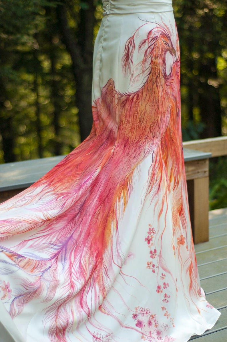 May include: A floor-length white skirt featuring a hand-painted phoenix design in shades of red, orange, and pink. The artwork depicts the bird with flowing, detailed feathers that extend down the fabric, accented by small floral motifs near the hem.