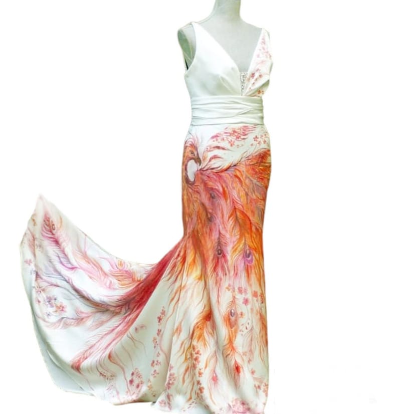 May include: A floor-length white sleeveless dress featuring a V-neckline and a wide waistband. The skirt displays a vibrant, hand-painted phoenix motif in shades of orange, red, and pink, with intricate feather details extending into a train.