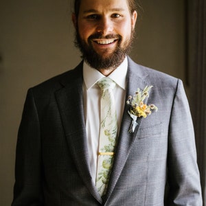 Puede incluir: Un hombre con un traje gris, camisa blanca y corbata con estampado floral. Lleva un boutonniere con flores amarillas y blancas.