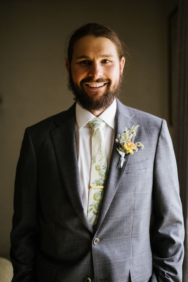 Puede incluir: Un hombre con un traje gris, camisa blanca y corbata con estampado floral. Lleva un boutonniere con flores amarillas y blancas.