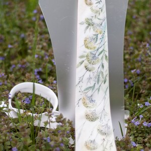 Puede incluir: Una corbata blanca con un patr&oacute;n floral de hojas verdes y flores blancas. La corbata se muestra en un soporte blanco sobre un fondo de hierba verde y flores moradas.