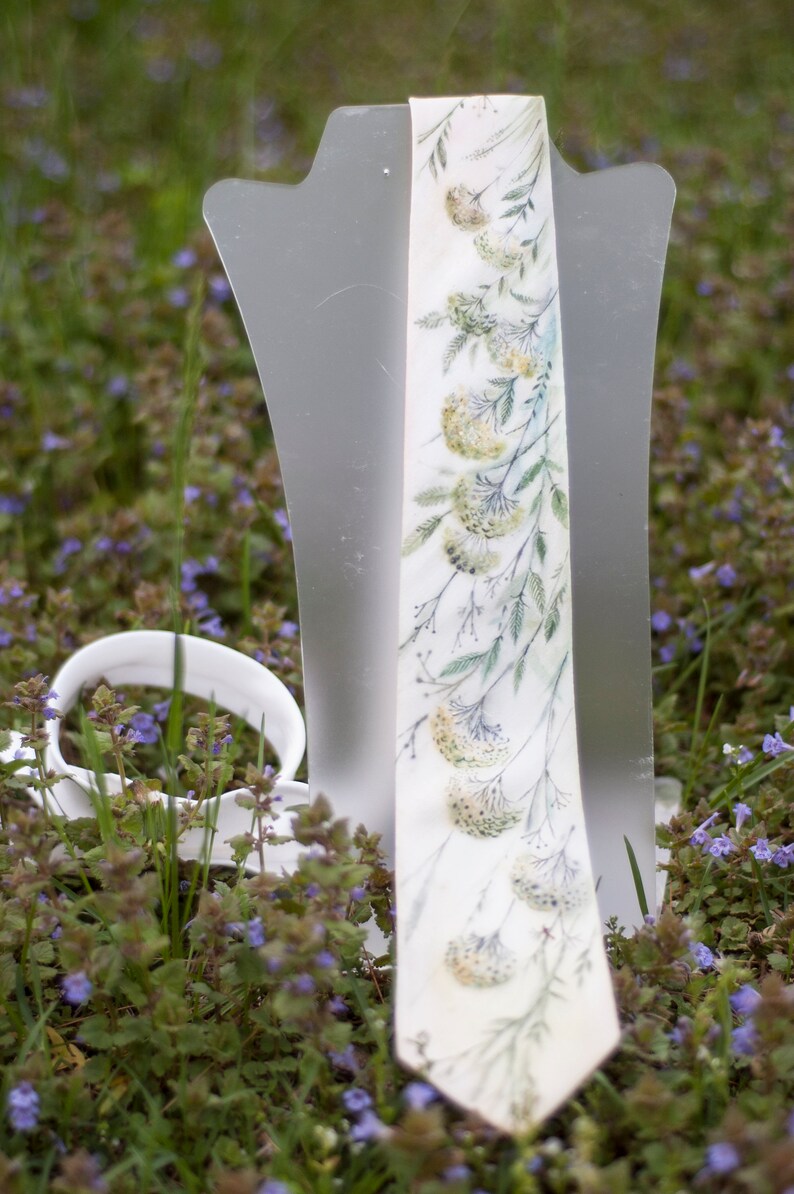 Puede incluir: Una corbata blanca con un patr&oacute;n floral de hojas verdes y flores blancas. La corbata se muestra en un soporte blanco sobre un fondo de hierba verde y flores moradas.