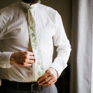 Puede incluir: Una camisa blanca con una corbata de estampado floral verde y blanco. La corbata est&aacute; siendo ajustada por una mano.