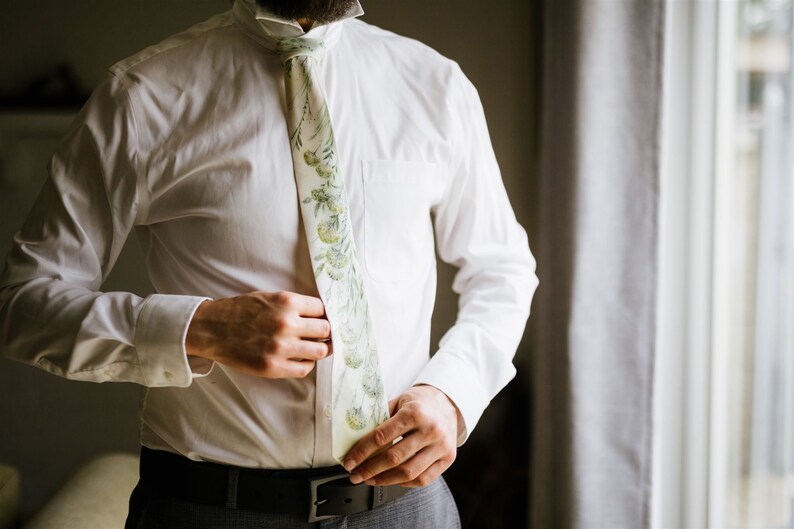 Puede incluir: Una camisa blanca con una corbata de estampado floral verde y blanco. La corbata est&aacute; siendo ajustada por una mano.