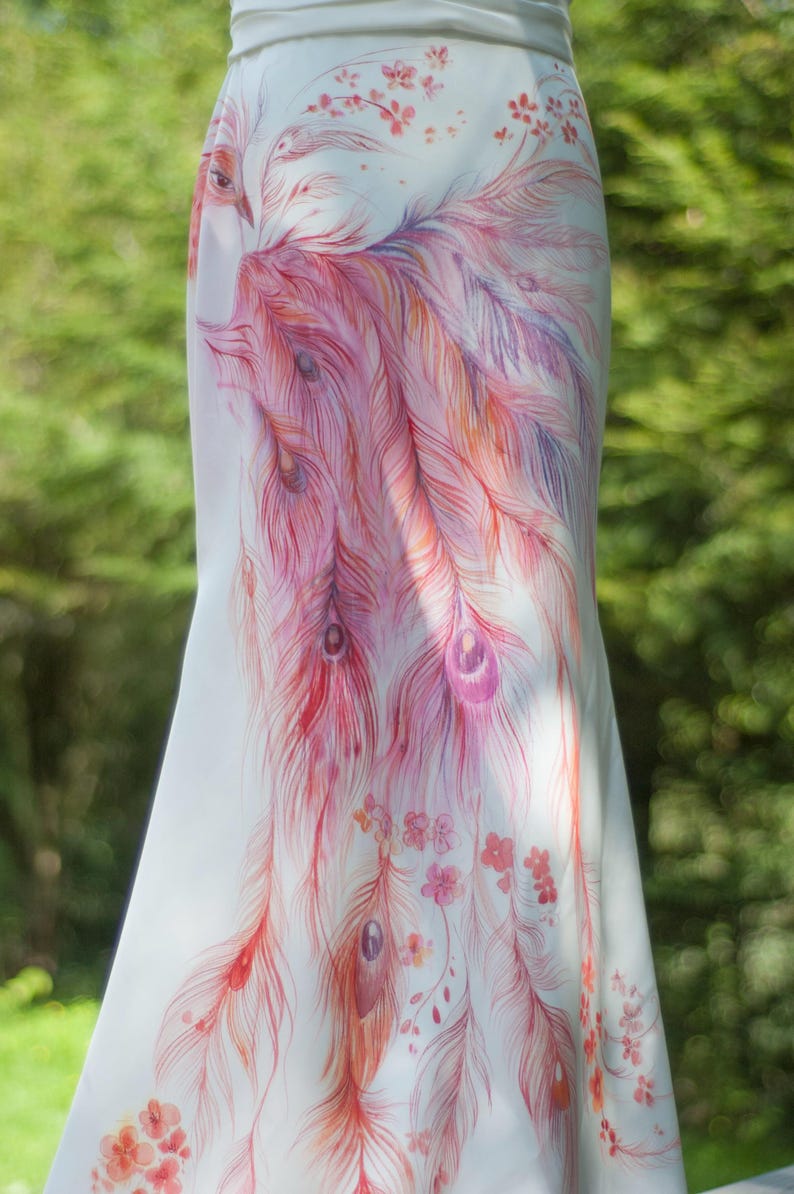 May include: A floor-length white skirt featuring a detailed hand-painted design of a large bird with long, flowing tail feathers in shades of pink, orange, and purple, accented with small floral motifs scattered across the fabric.