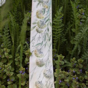 Puede incluir: Una corbata de seda blanca con un patr&oacute;n floral de hojas verdes y flores amarillas. La corbata est&aacute; sobre un lecho de plantas verdes.